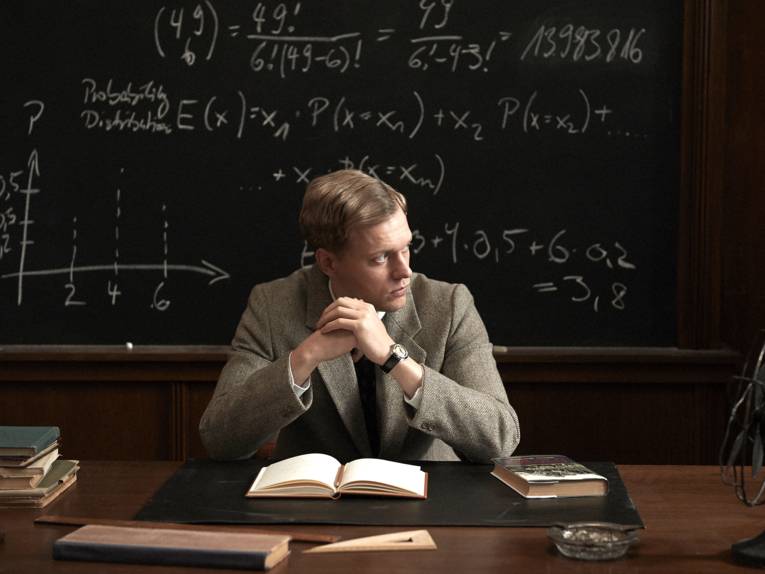 Ein Mann sitzt an einem Schreibtisch. Vor ihm ein aufgeschlagenes Buch, hinter ihm eine Tafel mit ma-thematischen Formeln